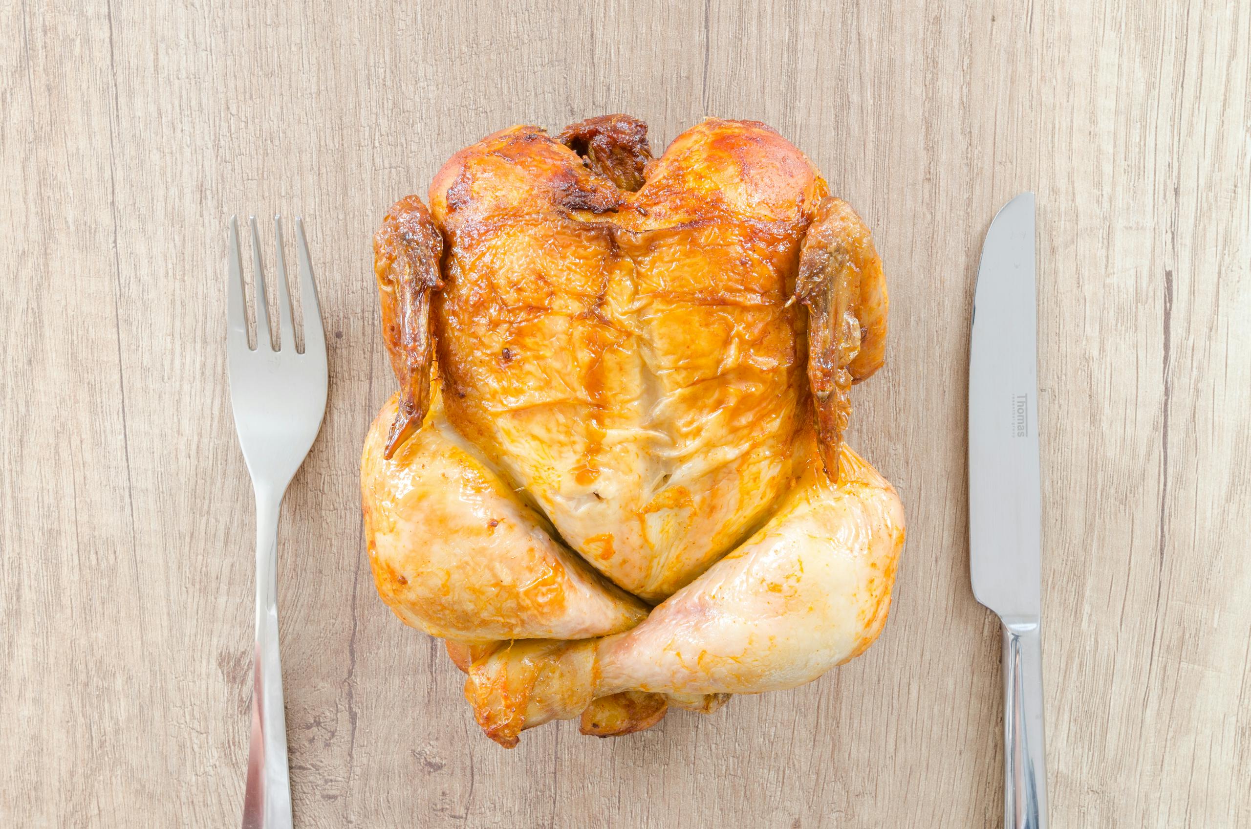 Top view of a perfectly roasted chicken placed between fork and knife on a wooden table.