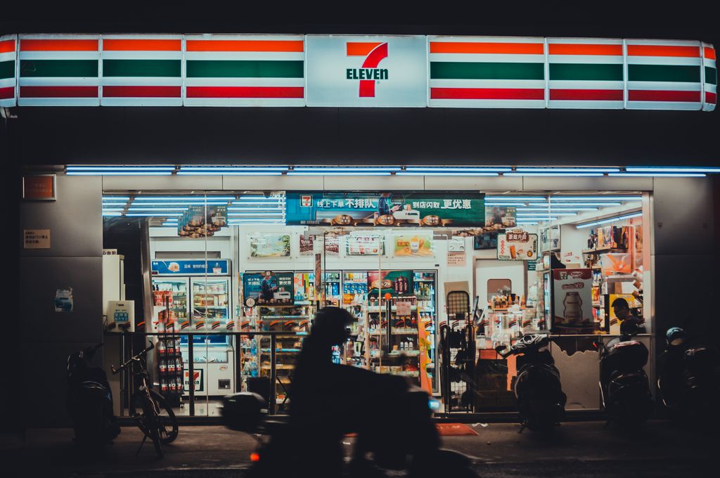A vibrant 7-Eleven store front lit up during nighttime with a silhouette of a motorbike rider passing by.