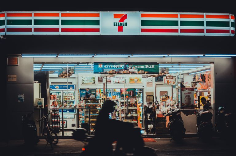 A vibrant 7-Eleven store front lit up during nighttime with a silhouette of a motorbike rider passing by.