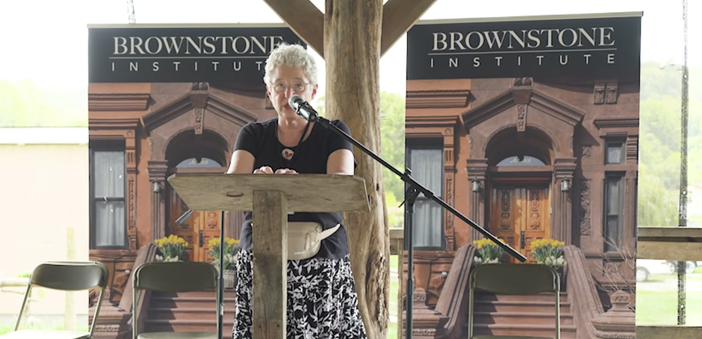 Meryl Nass stands at a podium with Brownstone Institute banners behind her