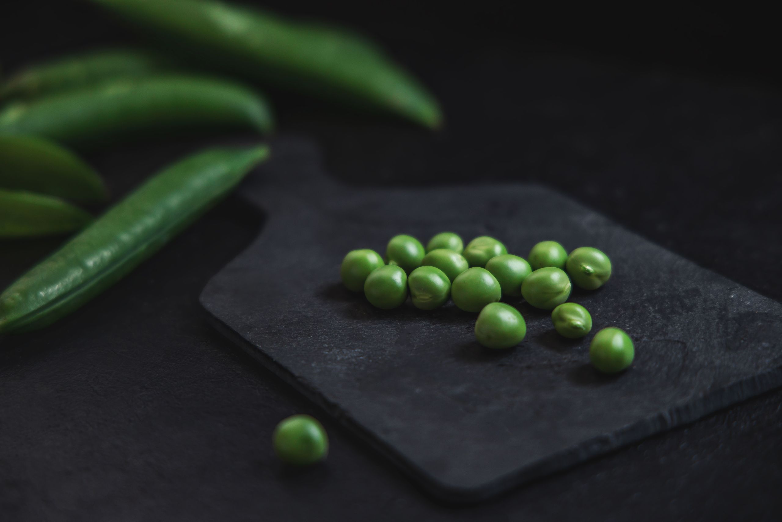 Vibrant green peas arranged on a black slate board with pea pods in background.